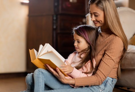 Profile Side View Portait Of Happy Smiling Mother And Cute Little Daughter Reading Book Together Sitting On Floor Leaning Couch At Home, Spending Time Together. Bedtime Story Concept