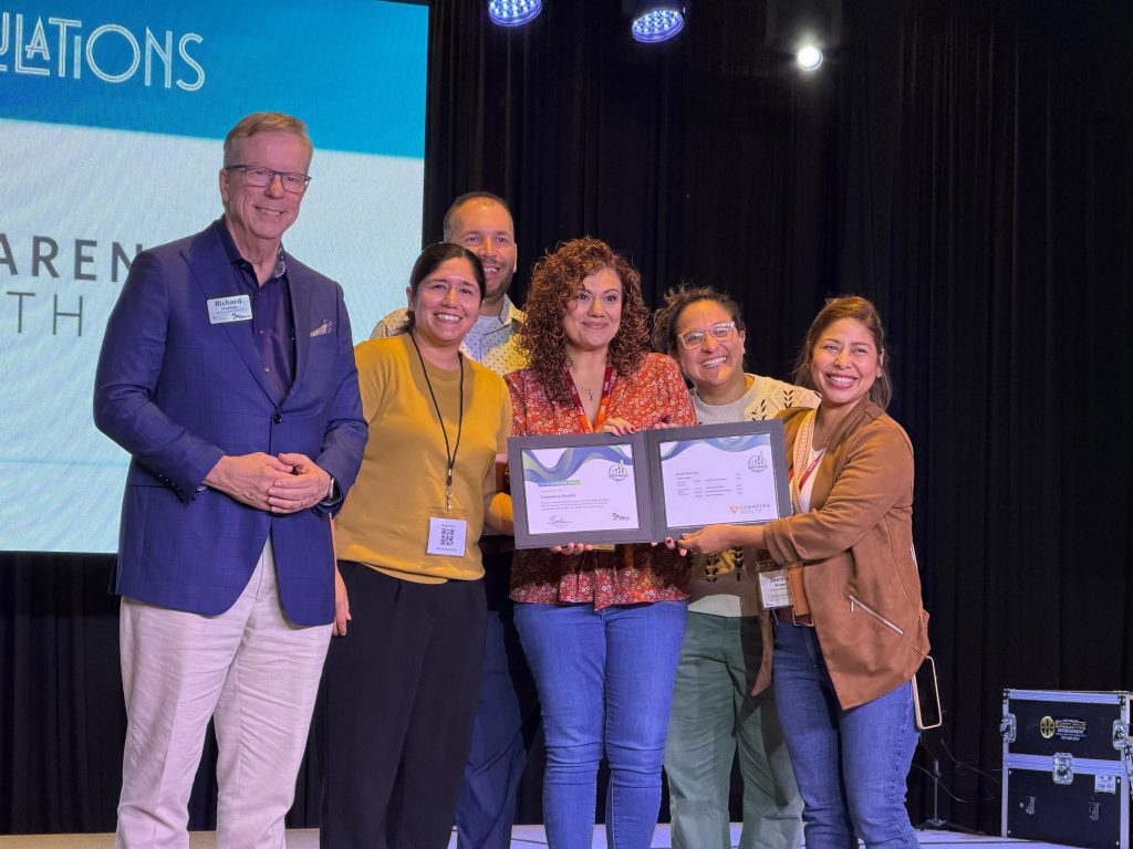 Six adults stand on a stage smiling; one proudly holds two certificates. Behind them, a screen displays part of "CONGRATULATIONS." The group appears to be celebrating a Camarena Health achievement or award.