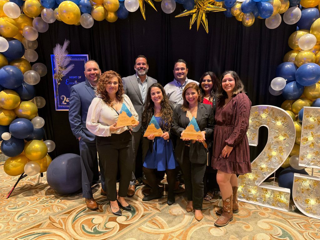 Eight people pose and smile in front of a balloon arch decorated with blue, gold, and white balloons at a Camarena Health event. Three women in front hold wooden awards, and a large “25” sign is decorated with lights to the right.