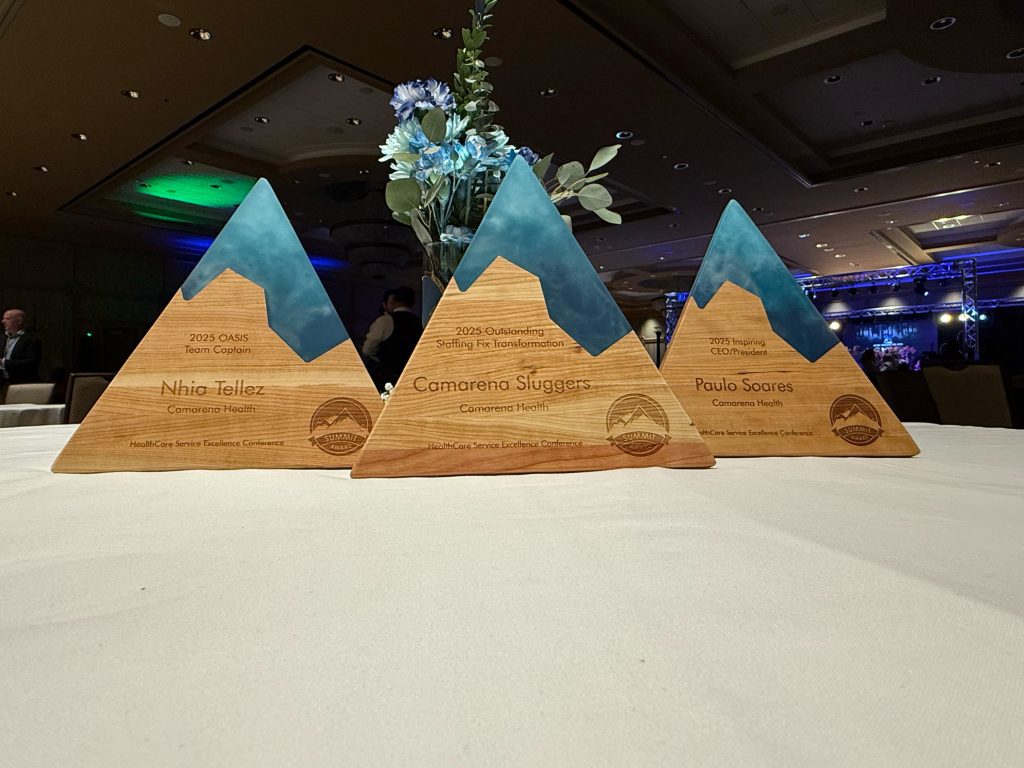 Three wooden, mountain-shaped Camarena Health awards with blue tops are displayed on a table, each engraved with recipients’ names and achievements, alongside a flower arrangement and a blurred event background.