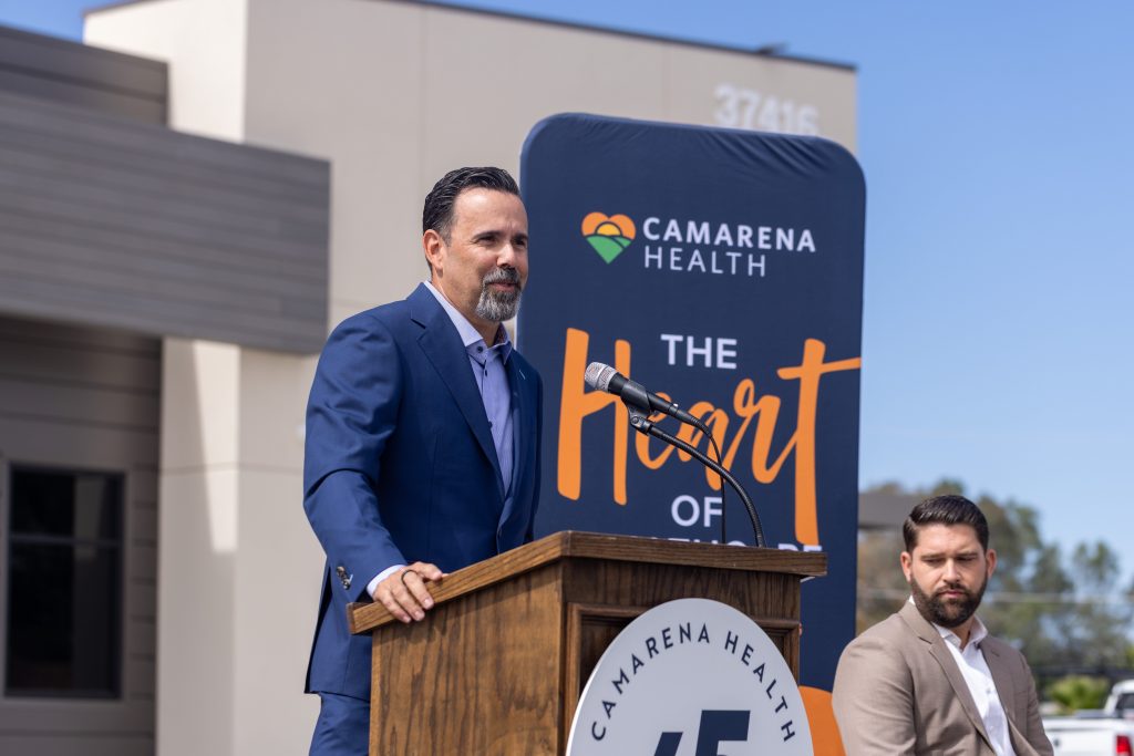 A man in a blue suit speaks at a podium during an outdoor event for Camarena Health, with another man seated beside him and a backdrop displaying the Camarena Health logo and the words The Heart Of.