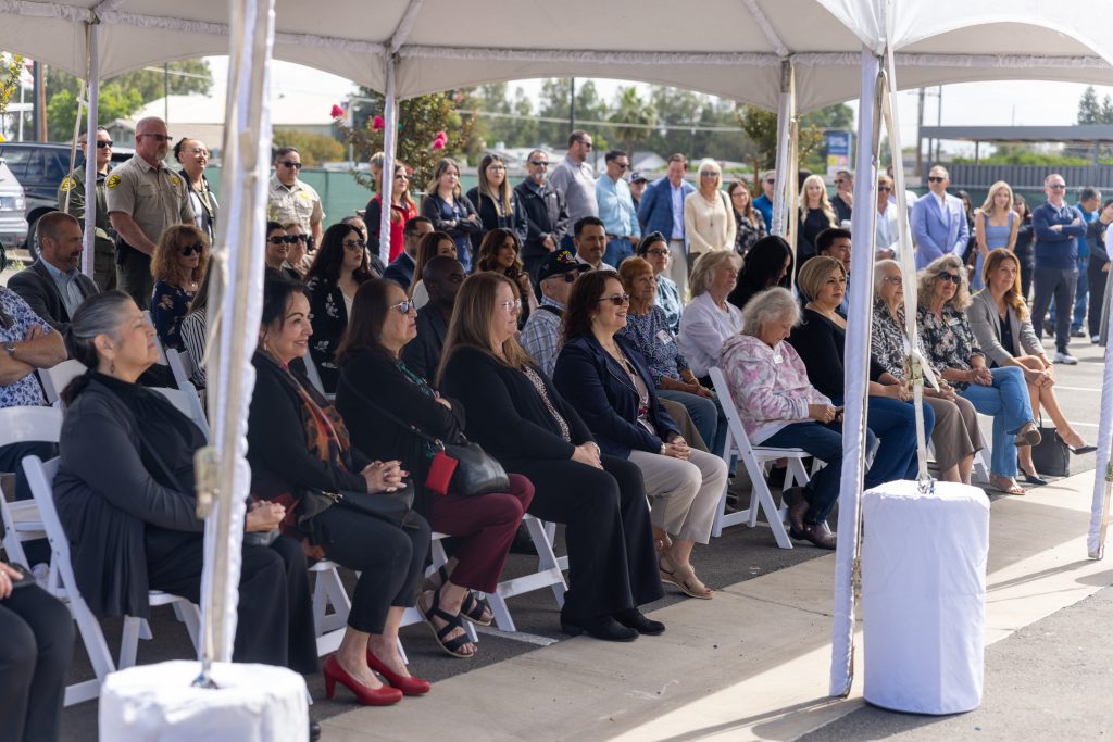A group of people sit under a white tent, attentively watching an outdoor event. Some are seated in folding chairs, while others stand in the background. The weather appears sunny and clear.