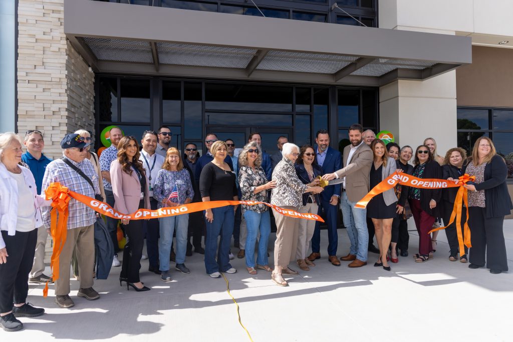 A large group of people stand in front of a building, smiling and holding a large orange Grand Opening ribbon as several people cut it to celebrate the opening event.