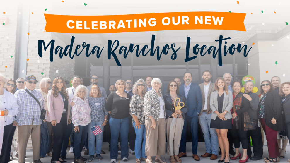 Ranchos Ribbon Cutting-blog A large group of people stands smiling in front of a building under a banner that reads, Celebrating Our New Madera Ranchos Location. Confetti is falling in the background.