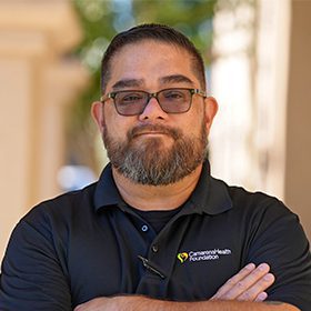 A man with a beard and glasses stands outdoors with his arms crossed, wearing a black polo shirt with a CommonSpirit Health Foundation logo. The background is blurred with bright natural light.