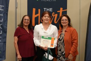 Three women stand indoors, smiling at the camera. The person in the center holds a certificate. A banner behind them reads The Heart with the word HEALTH above it. The two women on the sides wear glasses.