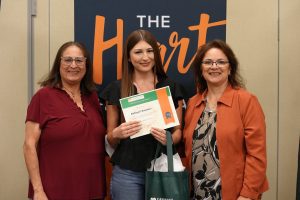 Three women stand together smiling; the woman in the center holds a certificate and a green bag. A sign behind them reads The Heart. They appear to be at a recognition or award event.