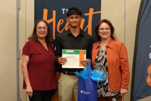 Three people stand indoors and smile at the camera. The young man in the center holds a certificate, while the women on each side stand close, one holding a blue gift bag. A banner with the word “Heart” is visible in the background.