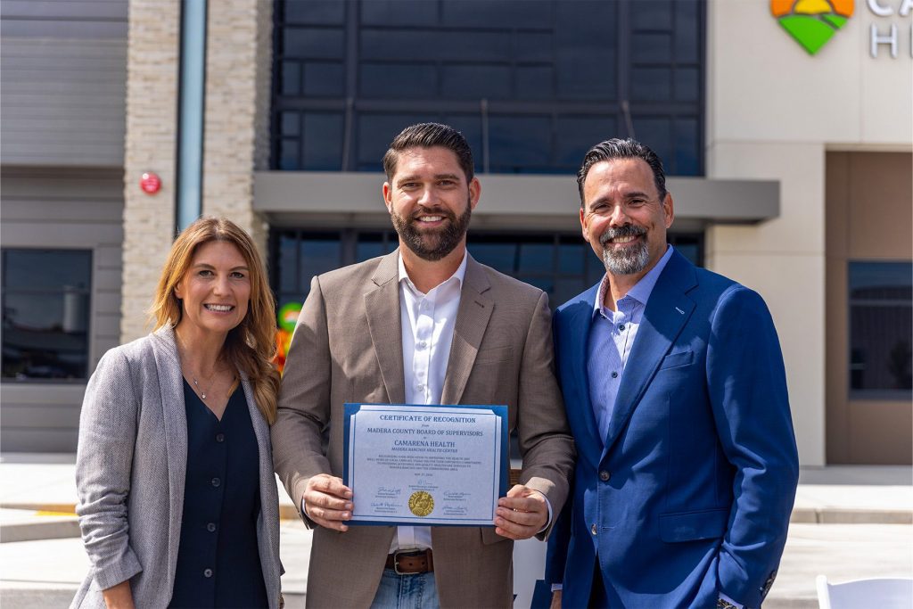 Three smiling people stand outdoors in front of a modern building. The man in the center holds a certificate, while the two others, a woman and a man, stand on either side of him.
